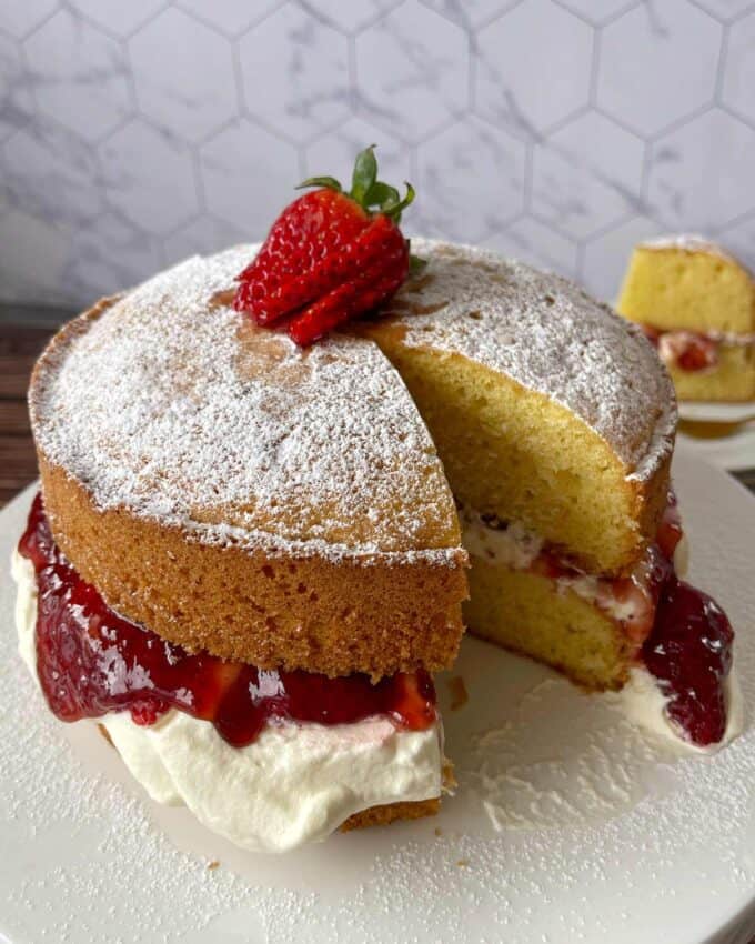 A layered sponge cake with whipped cream and strawberry filling, topped with a dusting of powdered sugar and a fresh strawberry, on a white plate.