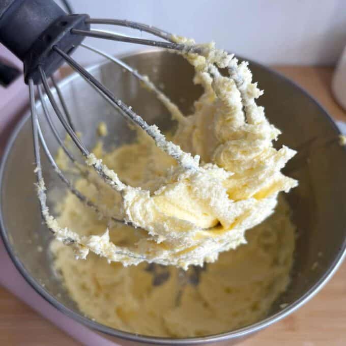 Butter and sugar mixture on a whisk attachment over a mixing bowl, indicating a baking process.