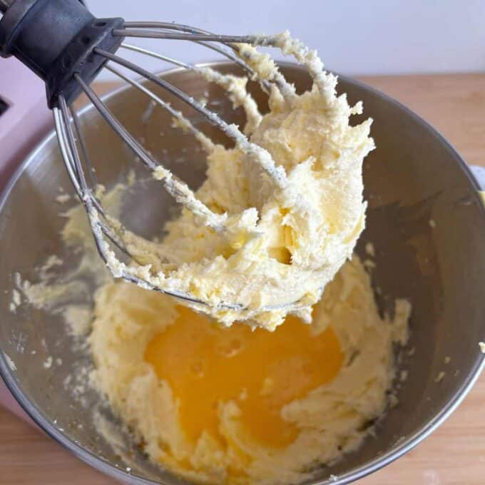 A mixing bowl with a whisk attachment covered in creamy batter. Melted butter is being added to the mixture below.