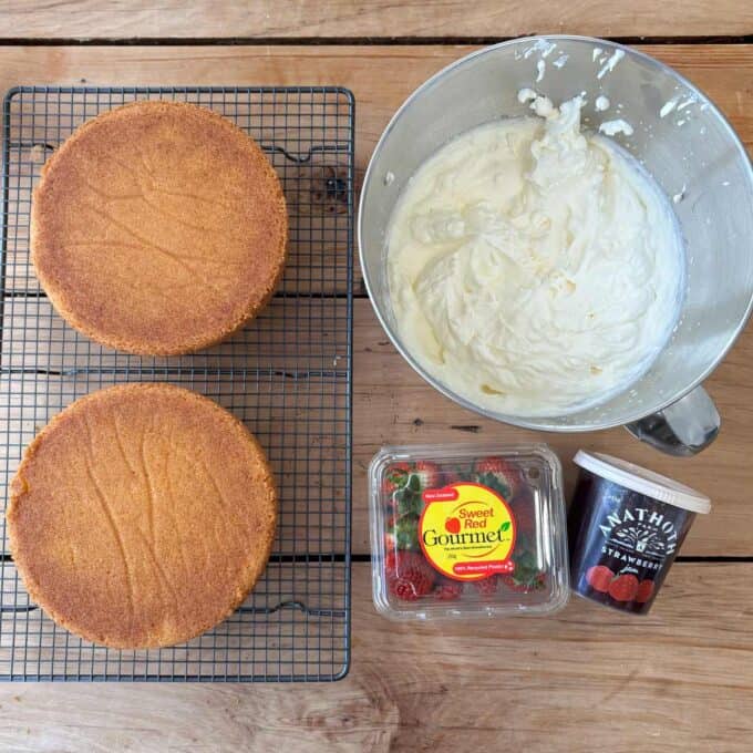 Two round sponge cakes on a cooling rack next to a mixing bowl with cream, a container of strawberries, and a jar of strawberry preserves on a wooden surface.