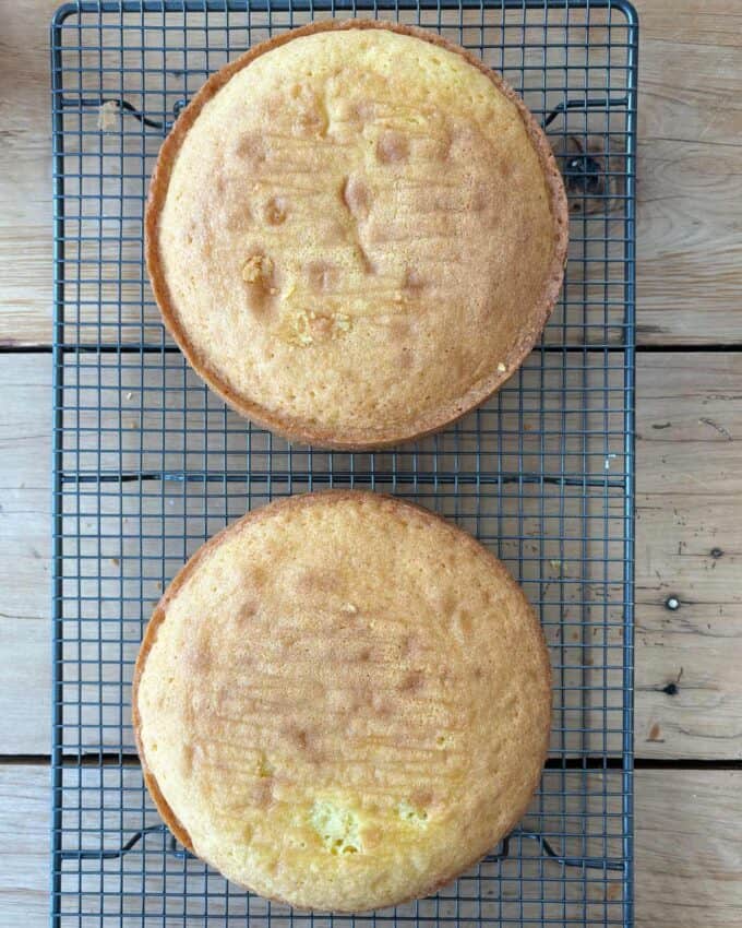 Two round yellow cakes are cooling on a wire rack placed on a wooden surface.