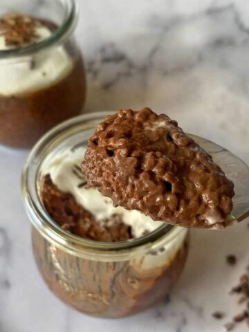 A spoonful of chocolate chia pudding is held above a glass jar filled with the same dessert, topped with whipped cream and chocolate shavings. Another jar is in the background.