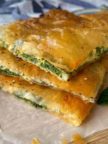Three stacked slices of spinach and cheese pie with golden, flaky pastry on parchment paper, garnished with mint leaves.