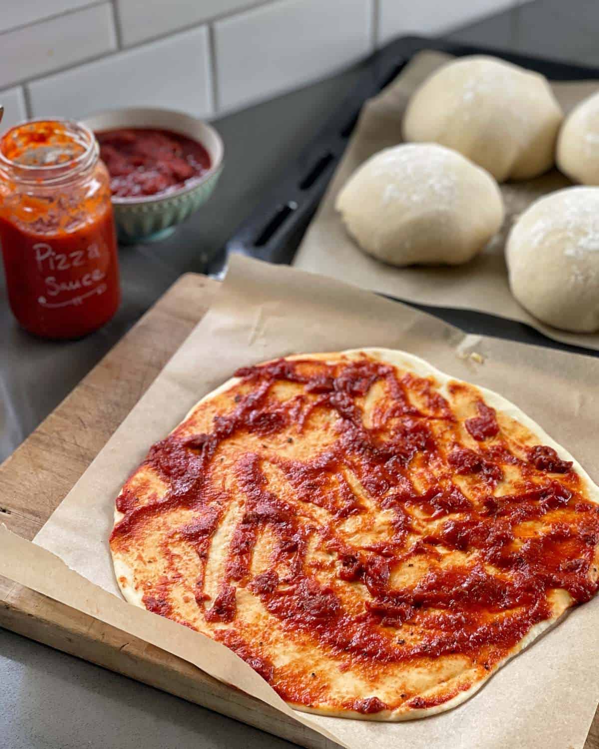 Homemade Pizza Sauce A rolled-out pizza dough spread with tomato sauce on parchment paper, with dough balls, a jar of pizza sauce, and a small bowl nearby.