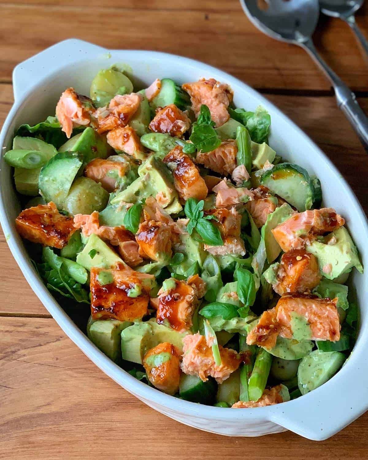 Salmon Potato Salad A white dish filled with grilled salmon and potato salad, featuring sliced cucumber, avocado, and leafy greens on a wooden table.