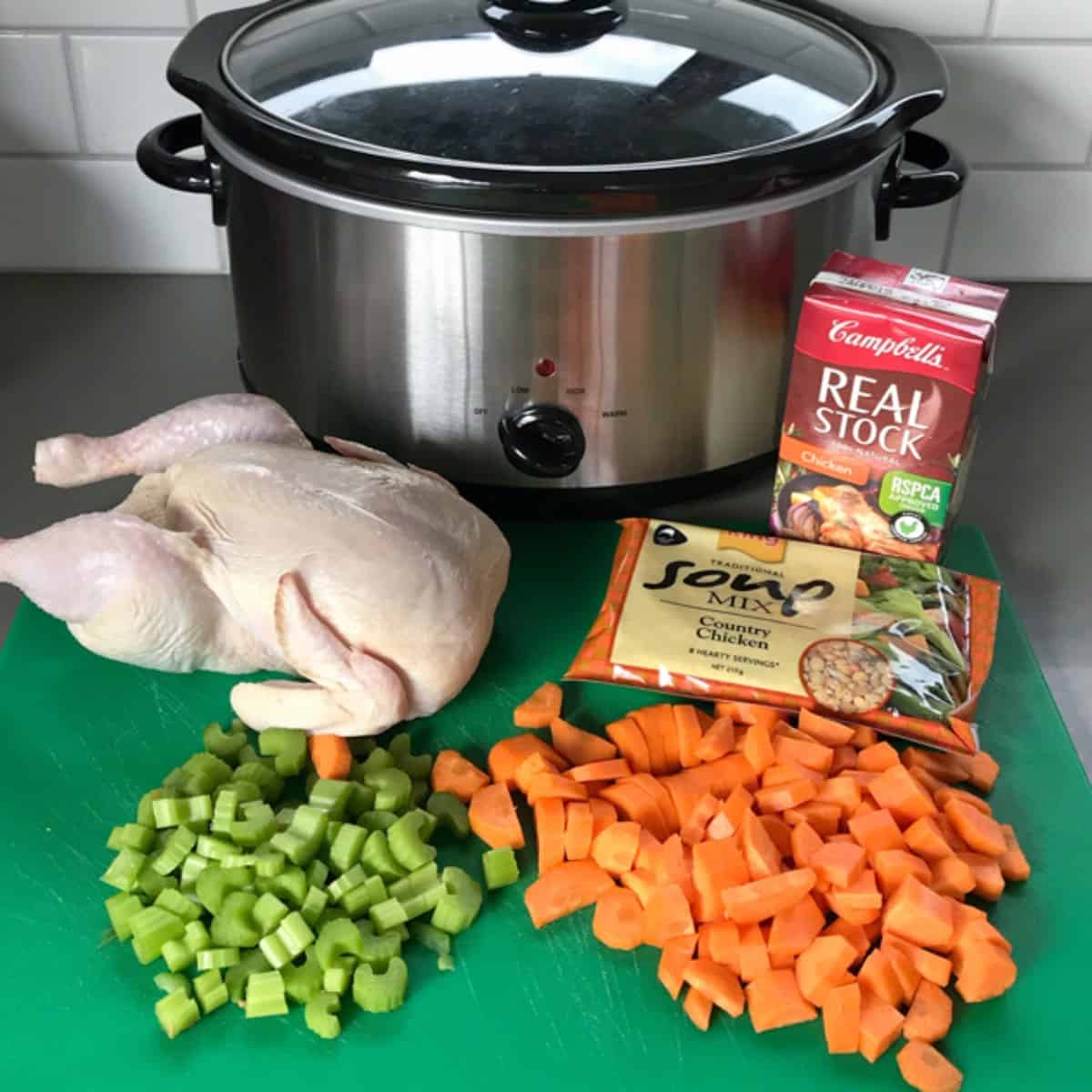 Slow Cooker Whole Chicken Soup Whole raw chicken, chopped carrots and celery, a packet of soup mix, chicken stock, and a slow cooker arranged on a countertop.