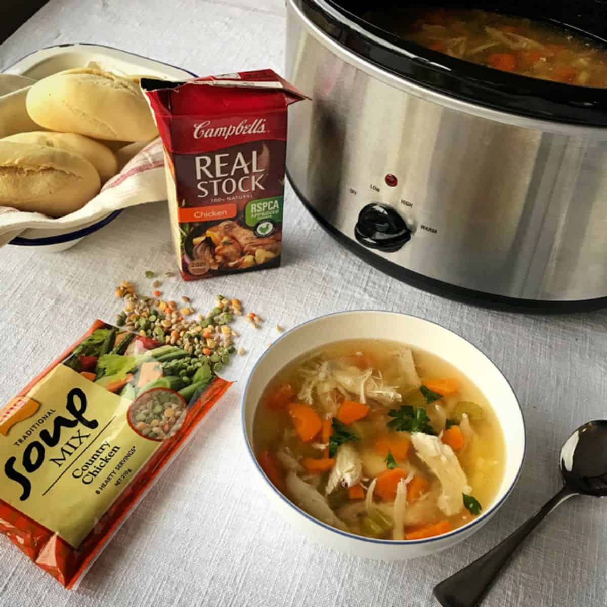 Slow Cooker Whole Chicken Soup A bowl of chicken and vegetable soup sits on a table next to a slow cooker, soup mix, chicken stock, fresh rolls, and a spoon.