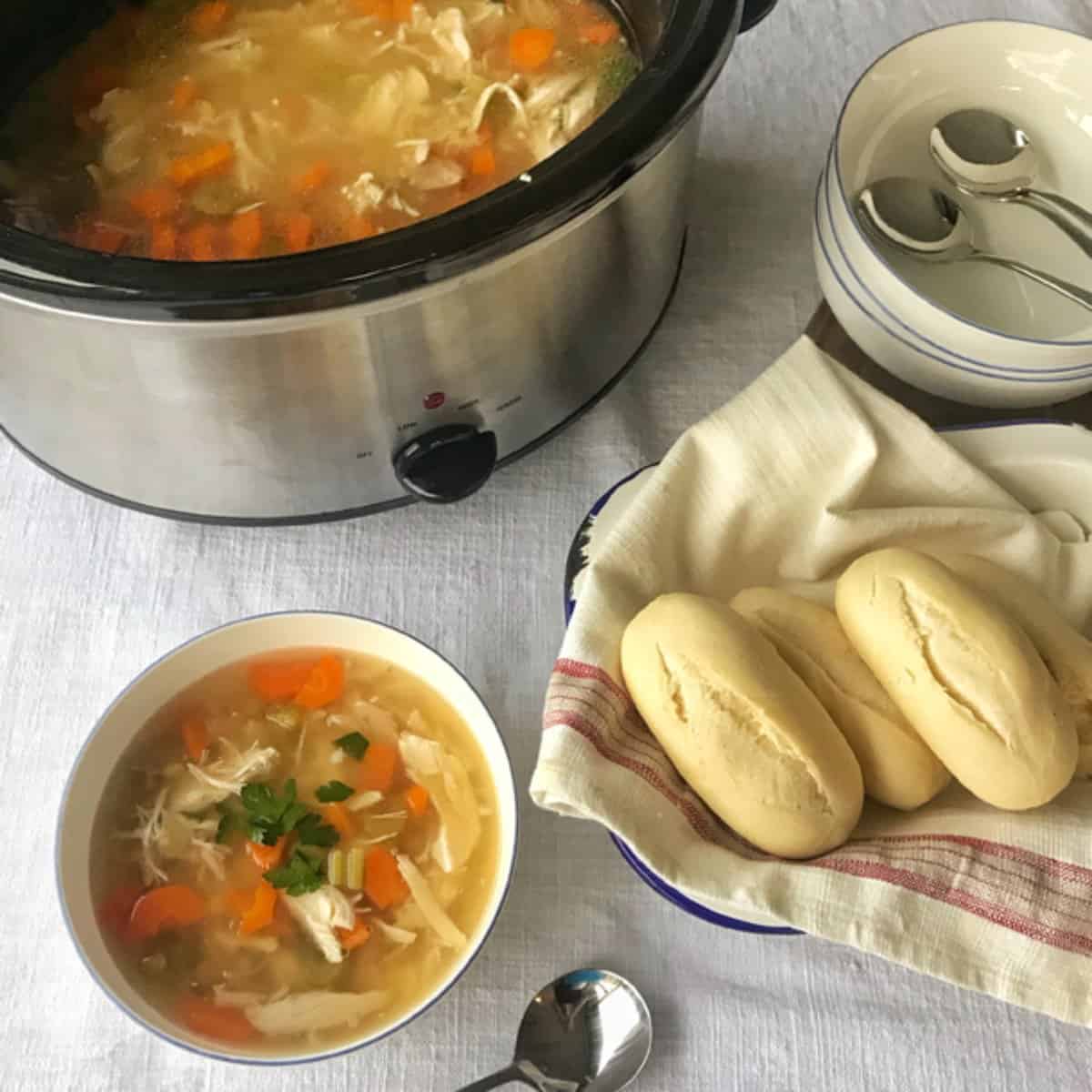 Slow Cooker Whole Chicken Soup A bowl of chicken soup with carrots and parsley sits next to a slow cooker, three bread rolls, stacked bowls, and spoons on a white tablecloth.