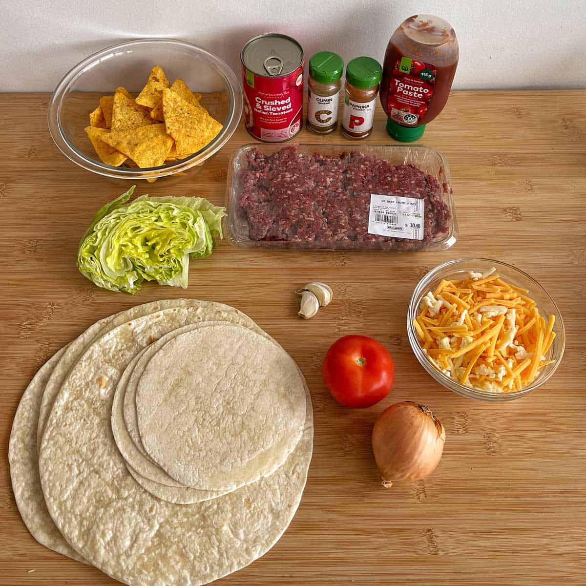 Ingredients for tacos on a wooden surface: tortillas, shredded lettuce, tomato, onion, garlic, ground beef, grated cheese, tortilla chips, canned tomatoes, spices, and tomato sauce.