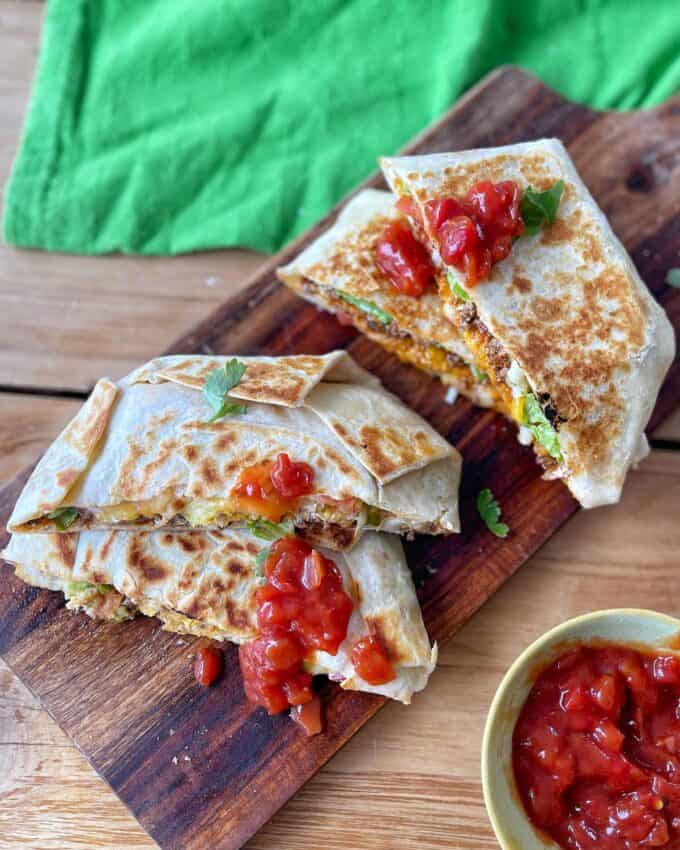 Two grilled burritos cut in halves on a wooden board, topped with salsa, with a small bowl of salsa nearby and a green cloth in the background.