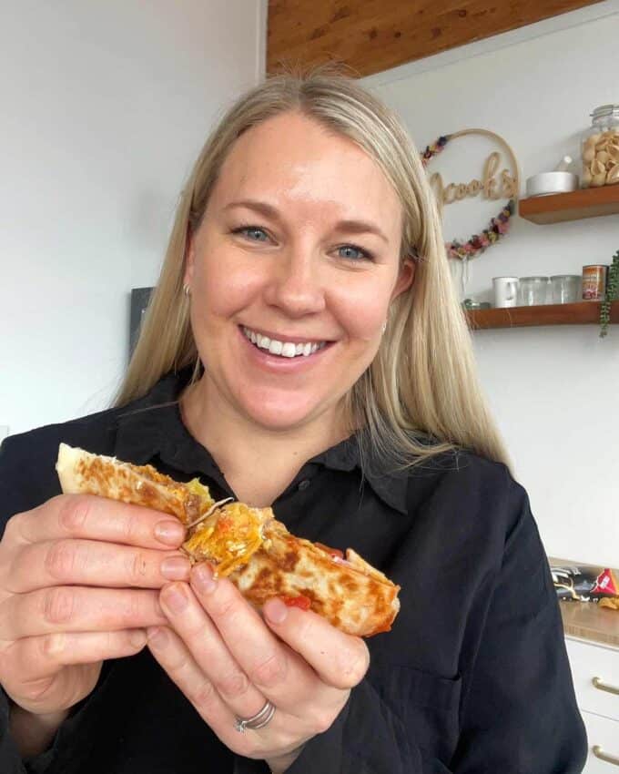 A woman smiling and holding a grilled wrap with a cheesy filling in a kitchen.