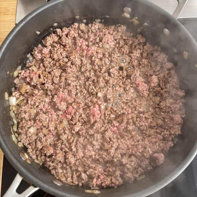 Ground beef and chopped onions cooking in a black skillet, with some steam rising from the pan.