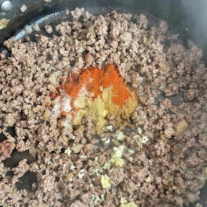 Ground beef cooking in a pan with chopped onions, minced garlic, and a pile of assorted spices including paprika, chili powder, cumin, and salt added on top.