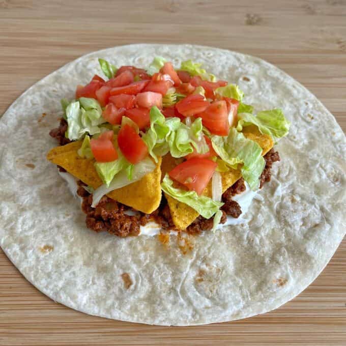 A flour tortilla topped with ground beef, sour cream, tortilla chips, shredded lettuce, and diced tomatoes on a wooden surface.