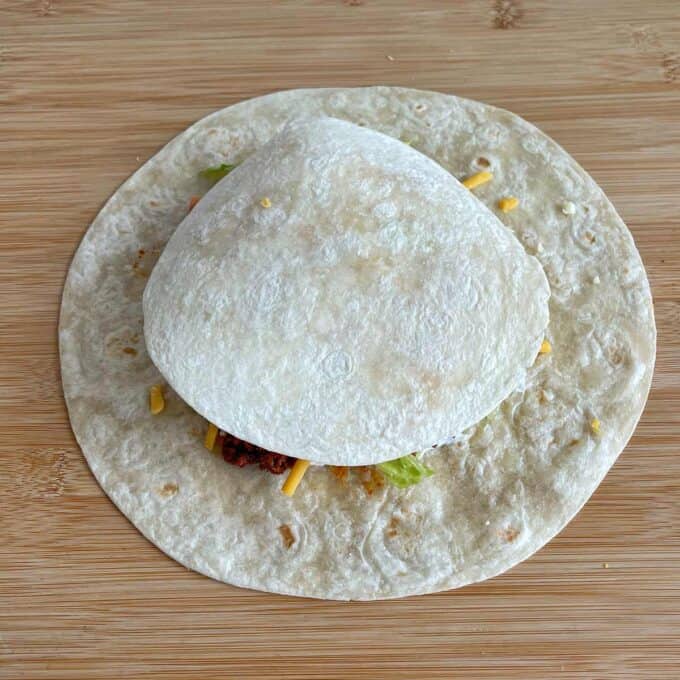 Two flour tortillas on a wooden surface, with the smaller one placed on top of taco fillings over a larger tortilla.