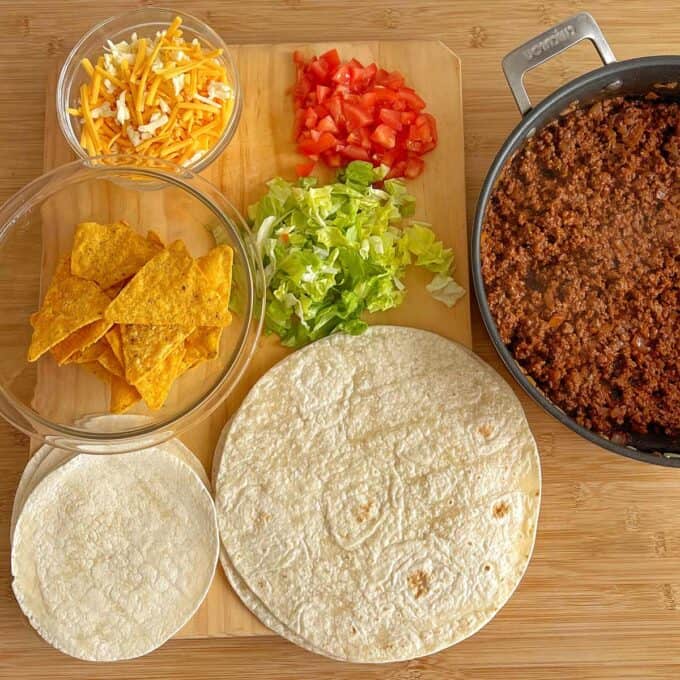 A cutting board with shredded cheese, diced tomatoes, chopped lettuce, tortilla chips, flour and corn tortillas, and a skillet of cooked ground beef on a wooden surface.