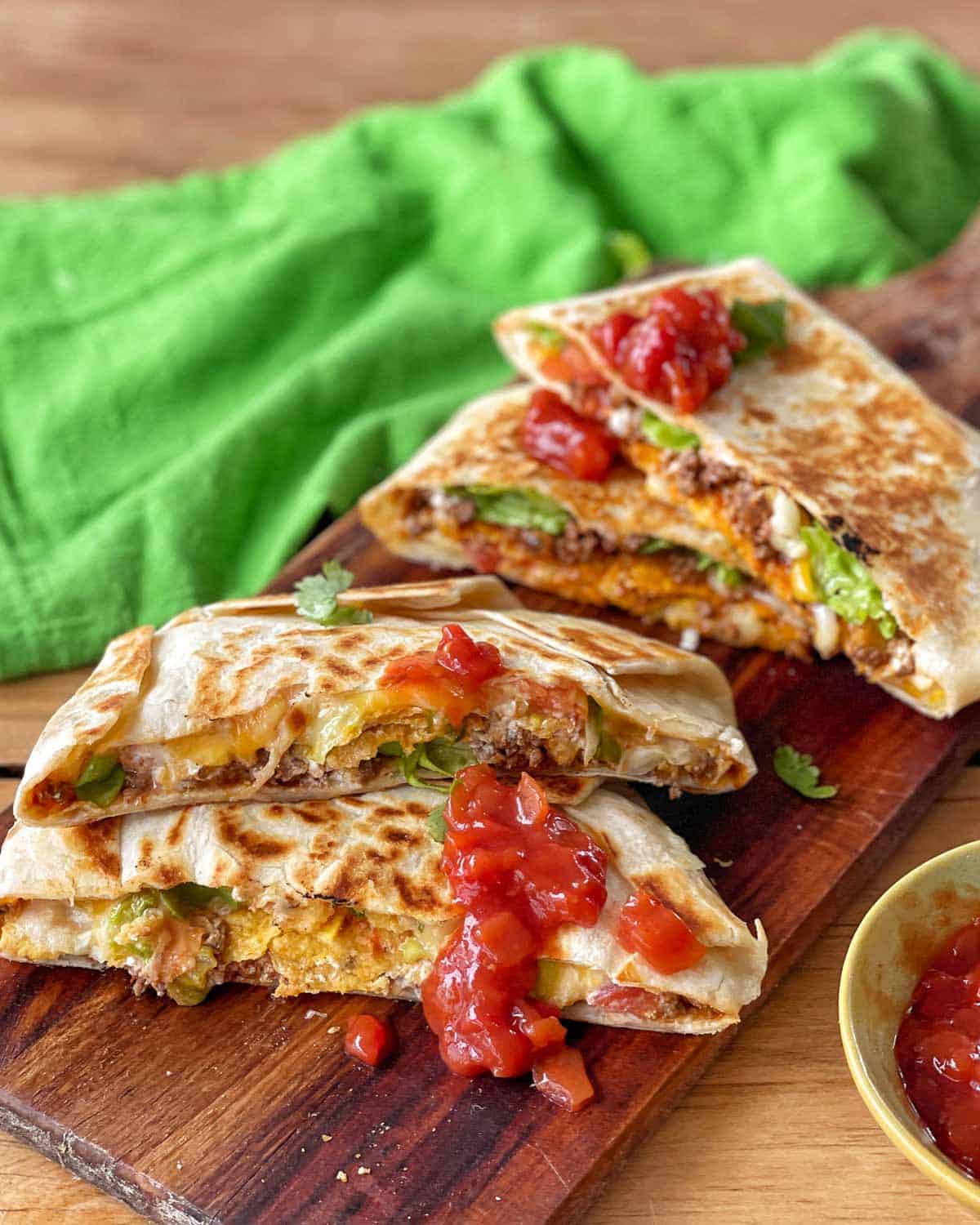 Beef Crunch Wraps Grilled tortilla wraps filled with ground meat, lettuce, and cheese, cut in half and topped with salsa, displayed on a wooden board with a green cloth in the background.