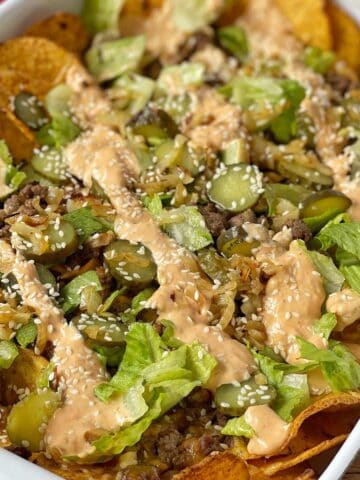A dish of nachos topped with ground beef, sliced pickles, shredded lettuce, sesame seeds, and a drizzle of creamy sauce.