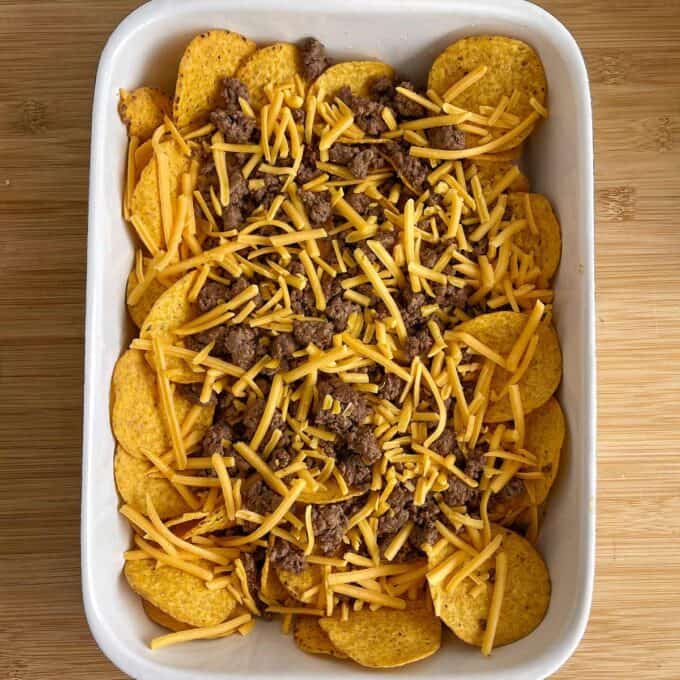 A white baking dish filled with corn chips, cooked ground beef, and shredded cheddar cheese on a wooden surface.