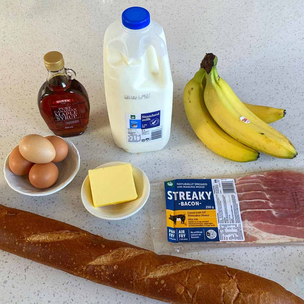 A loaf of bread, three eggs in a bowl, a bottle of maple syrup, a carton of milk, a block of butter, a bunch of bananas, and a pack of streaky bacon on a countertop.