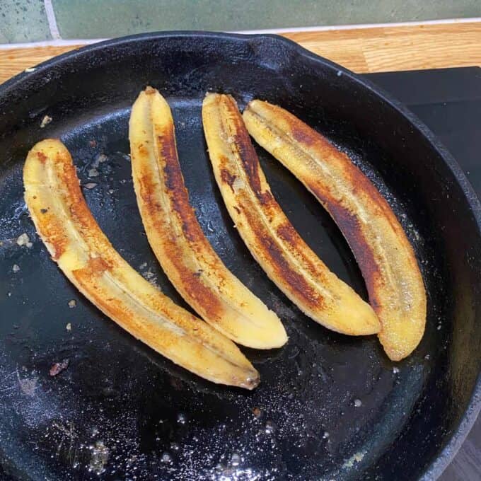 Four halved bananas being pan-fried in a black cast iron skillet on a stovetop, showing browning on the cut sides.