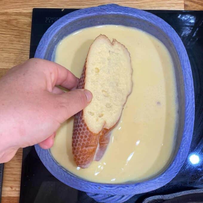 A hand dips a slice of bread into a blue dish filled with a pale yellow liquid, likely a batter or custard mixture.