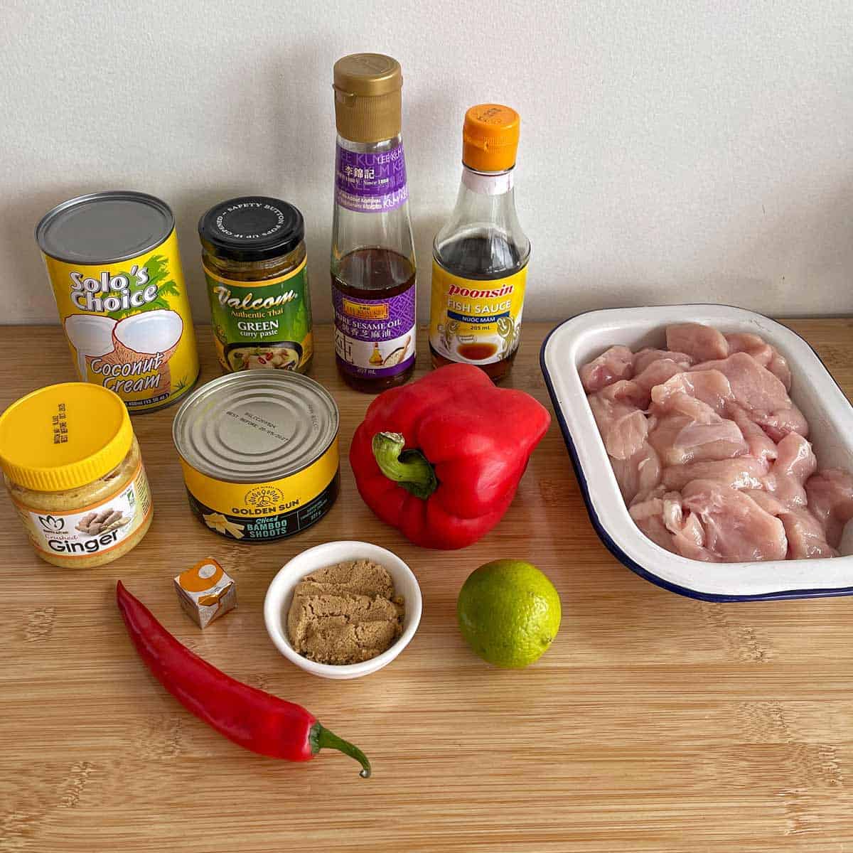 A selection of ingredients on a wooden surface, including raw chicken, red bell pepper, coconut cream, sauces, ginger, brown sugar, lime, chili, and canned bamboo shoots.