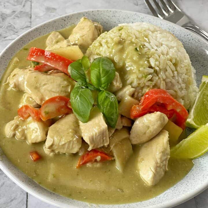 A plate of green curry chicken with red bell peppers and rice, garnished with fresh basil and lime wedges, with a fork and knife in the background.
