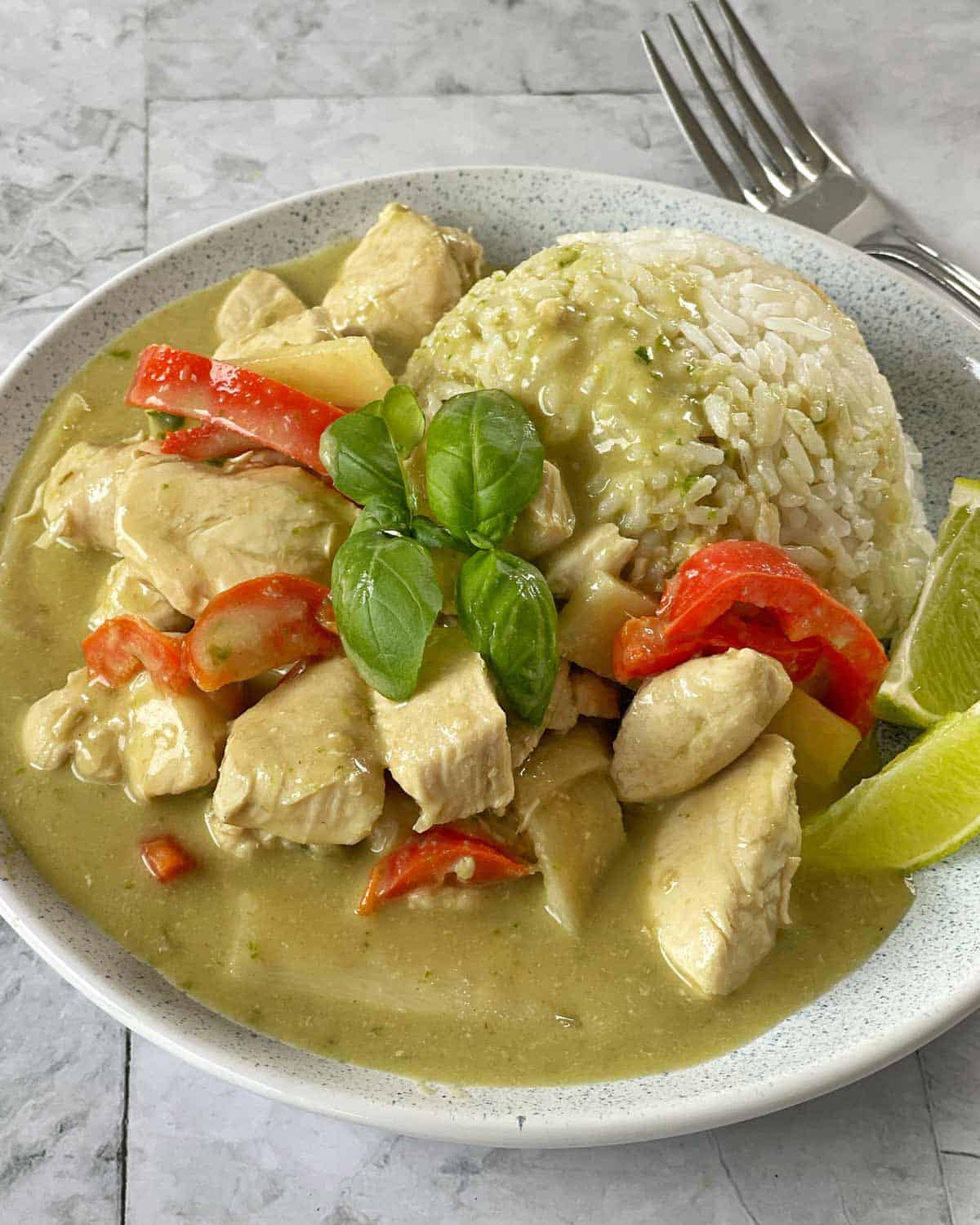 Chicken Thai Green Curry A plate of green curry chicken with red bell peppers and rice, garnished with fresh basil and lime wedges, with a fork and knife in the background.