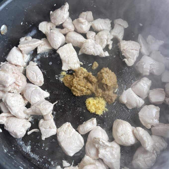 Cubed raw chicken pieces cooking in a skillet with minced garlic and a mound of brown seasoning paste in the center.