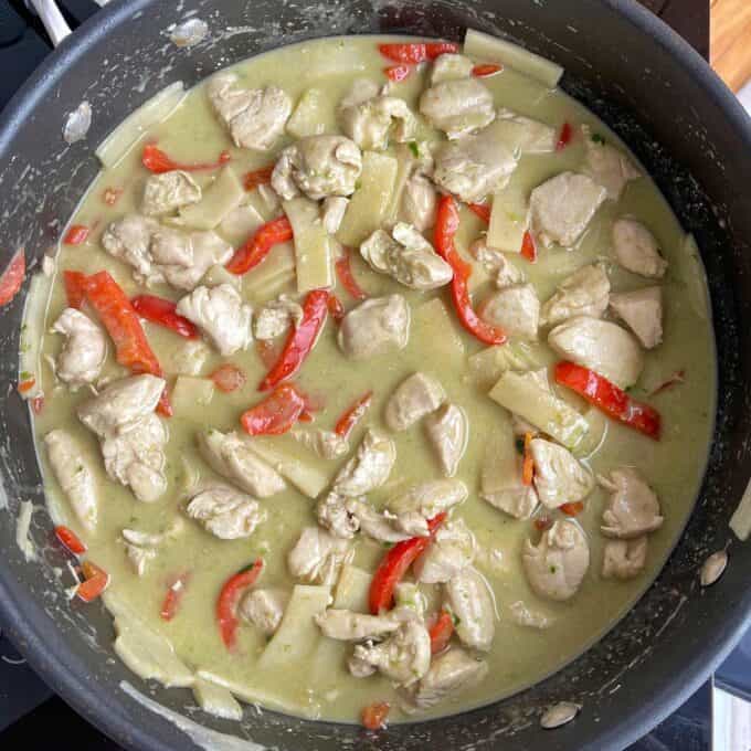 A pan filled with chicken pieces, red bell peppers, and bamboo shoots in a creamy green curry sauce.