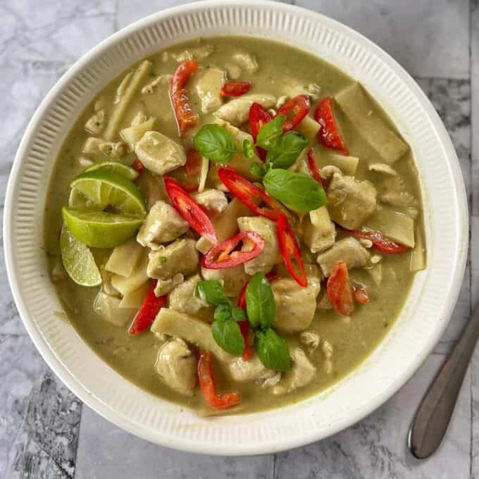 A bowl of Thai green curry with chicken, red bell peppers, bamboo shoots, fresh basil, sliced chili, and lime wedges, served on a marble surface with a spoon beside the bowl.