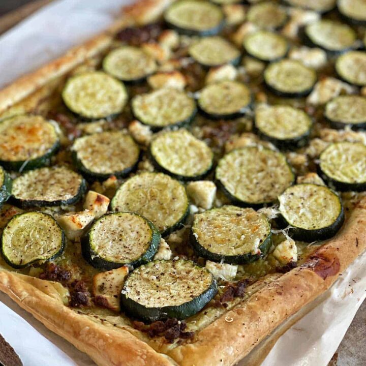 A rectangular puff pastry tart topped with sliced zucchini, crumbled cheese, and herbs, baked until golden brown on parchment paper.