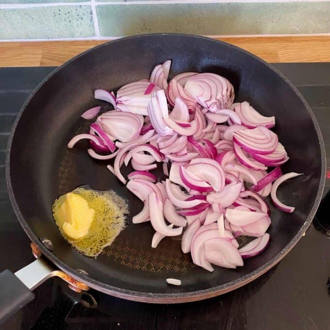 Sliced red onions and a pat of butter in a frying pan on a stovetop, with the butter starting to melt.