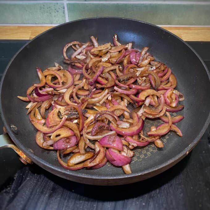 Sliced red onions sautéing in a black frying pan on a stovetop, beginning to caramelize.
