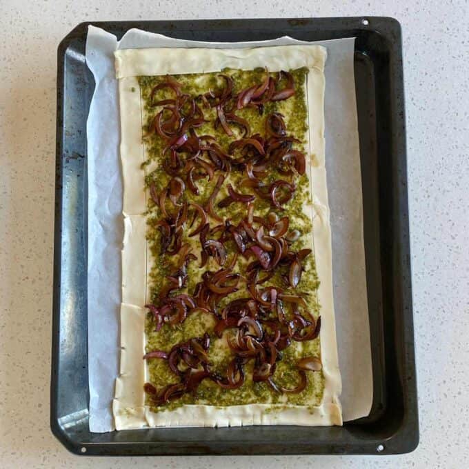A rectangular puff pastry topped with green pesto and caramelized red onions sits on parchment paper in a baking tray.