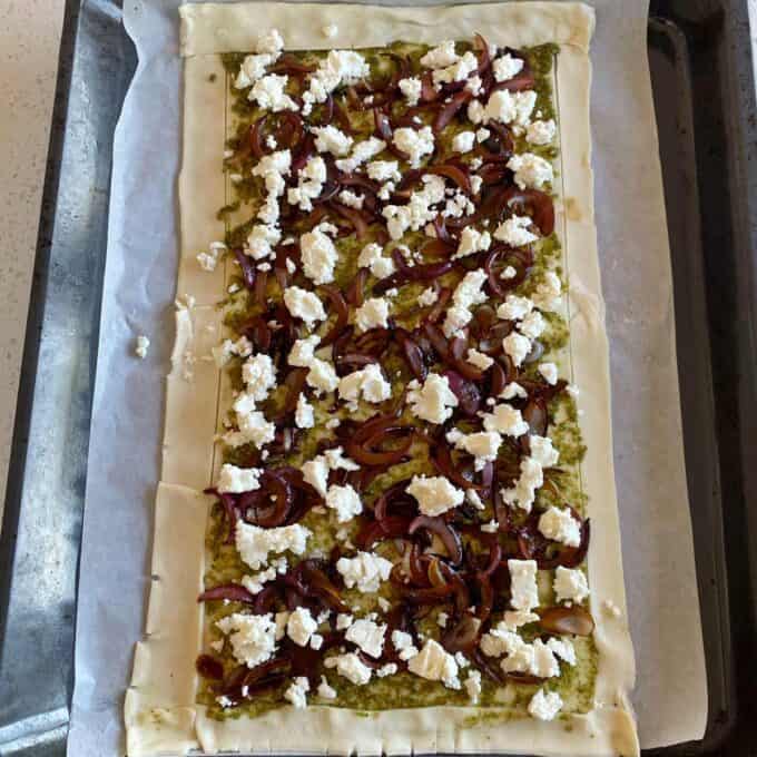 Unbaked rectangular pastry on parchment paper topped with pesto, caramelized onions, and crumbled feta cheese on a baking tray.
