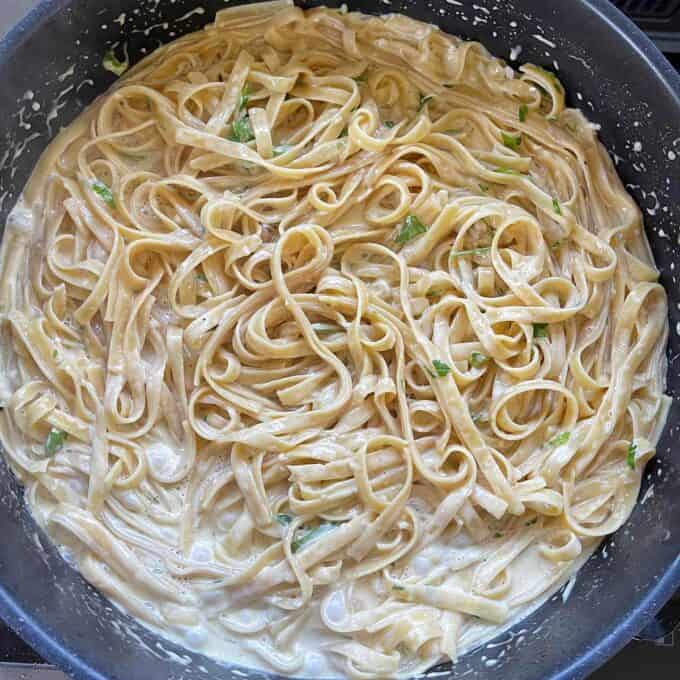A pan filled with cooked fettuccine noodles in a creamy white sauce, garnished with chopped green herbs.