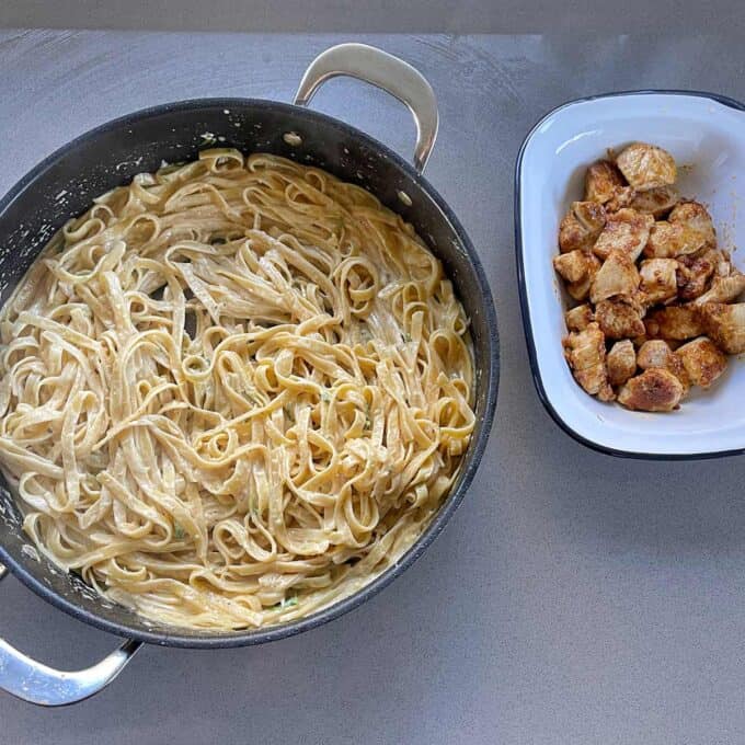 A pan of creamy fettuccine pasta sits next to a dish of cooked, seasoned chicken pieces on a gray countertop.