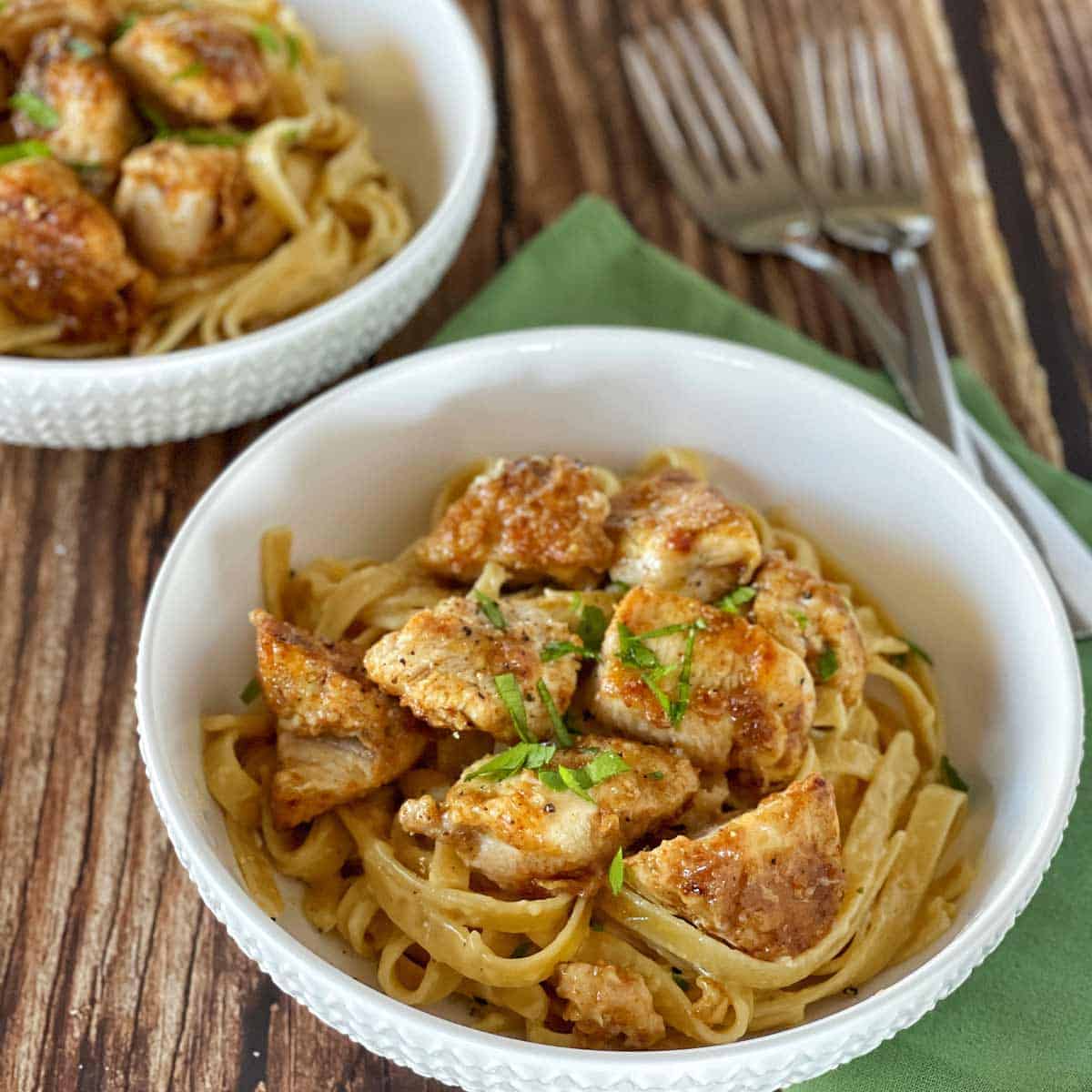 Two white bowls filled with fettuccine pasta and pieces of cooked chicken, garnished with herbs, placed on a wooden table with two forks and a green napkin nearby.
