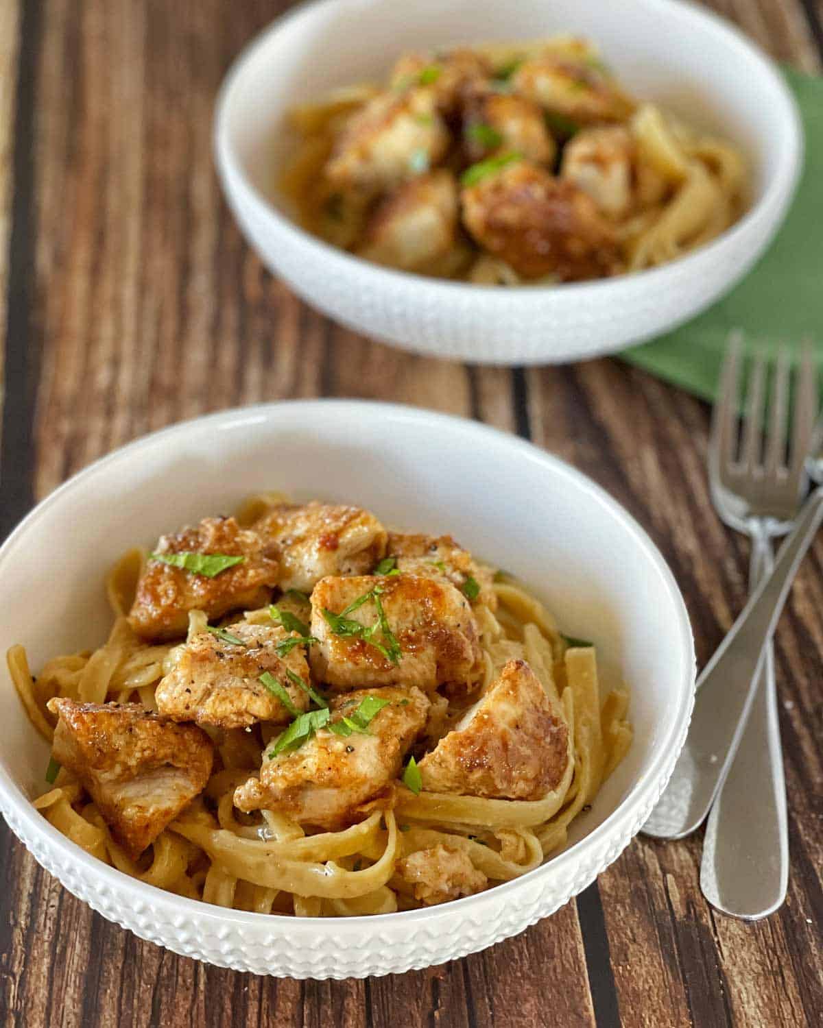 Creamy Fettuccine with Chicken Bites Two white bowls filled with fettuccine pasta and pieces of cooked chicken sit on a wooden table next to a fork and knife.