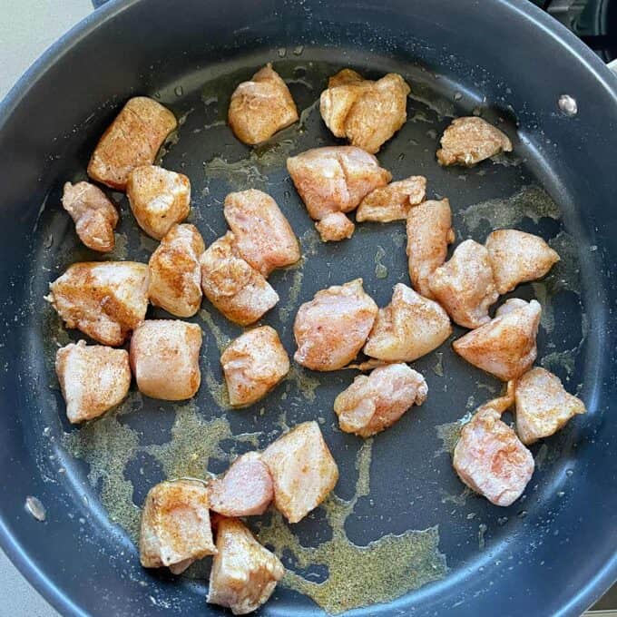 Raw, seasoned chicken pieces are spread out in a single layer on a black frying pan, ready to be cooked.