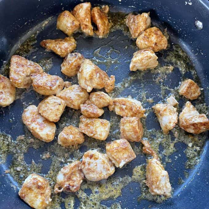Cubed pieces of chicken are being sautéed in a pan, showing browning and cooked juices in the pan.