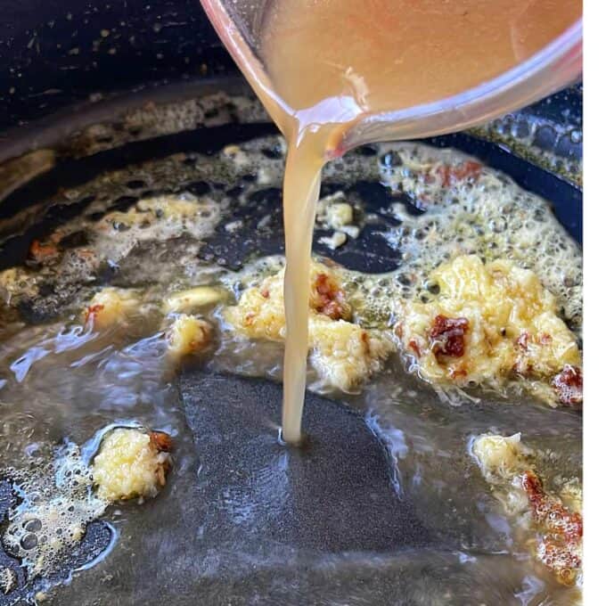 A close-up of broth being poured from a measuring cup into a pan with browned bits and bubbling fat.