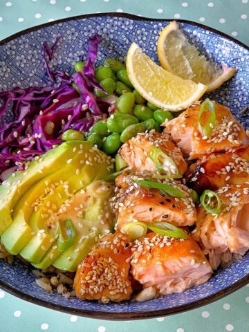 A blue bowl with cooked salmon, sliced avocado, shredded red cabbage, edamame, lemon wedges, green onions, and sesame seeds, on a green polka dot surface.