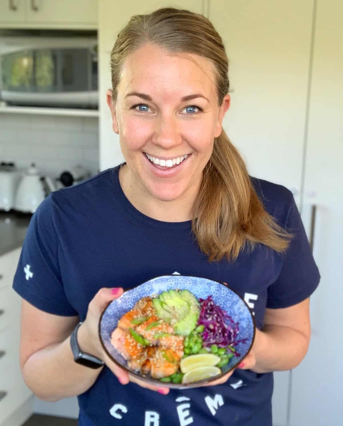 A woman stands in a kitchen, smiling and holding a bowl of colorful food topped with avocado, lime, and sesame seeds.