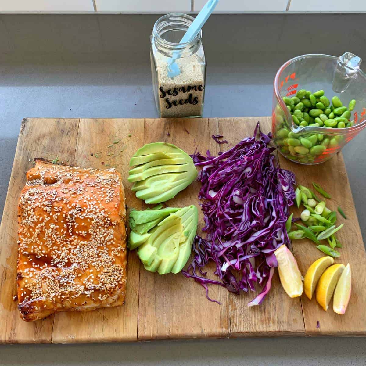 grilled honey soy salmon rice bowls A wooden board with cooked sesame-crusted salmon, sliced avocado, shredded red cabbage, green onions, lemon wedges, a cup of edamame, and a jar of sesame seeds.