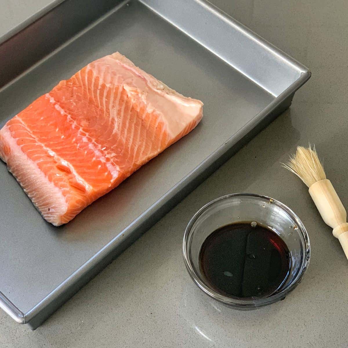 grilled honey soy salmon rice bowls A raw salmon fillet sits in a metal baking pan next to a small bowl of dark sauce and a basting brush on a gray countertop.