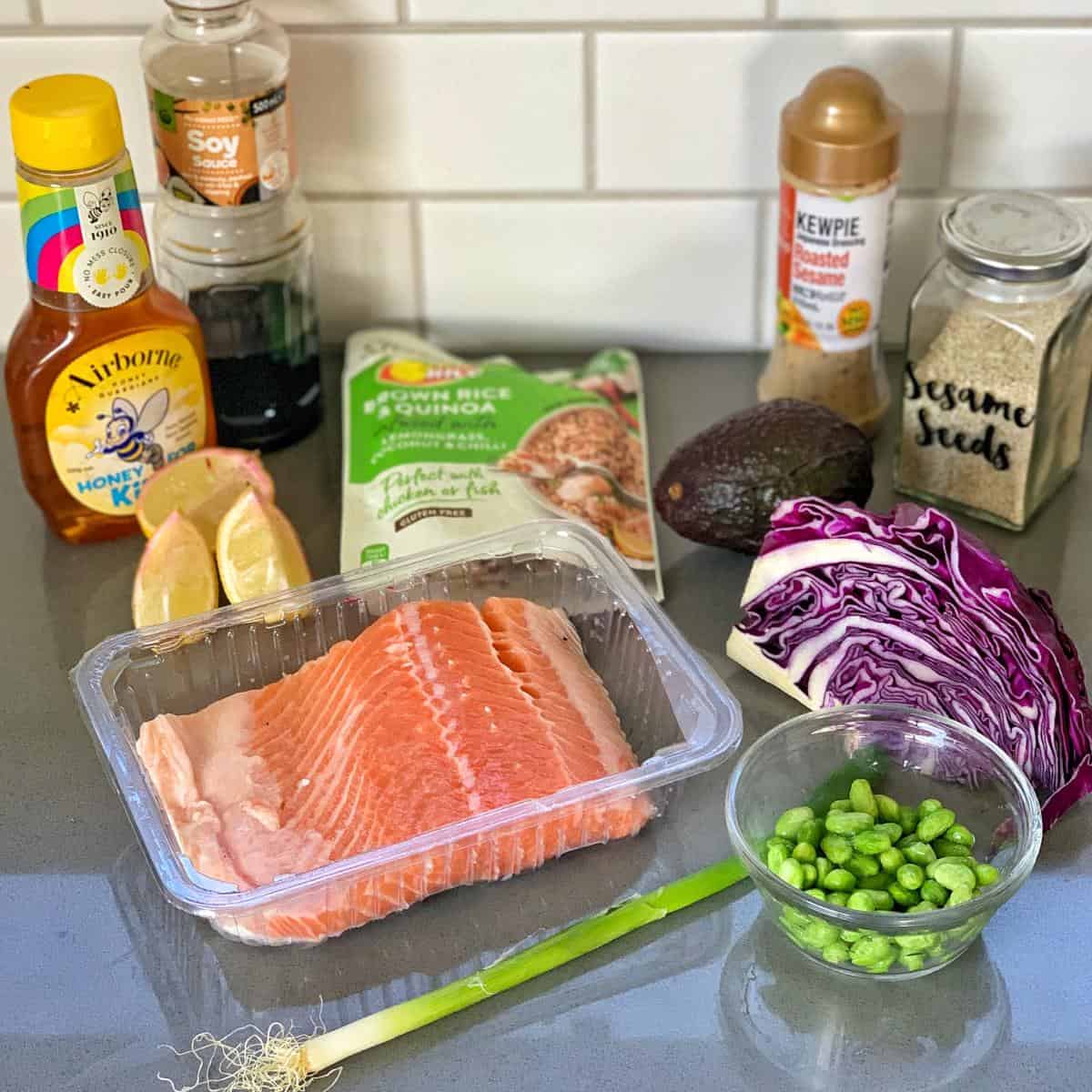 grilled honey soy salmon rice bowls Fresh salmon fillet, avocado, red cabbage, edamame, spring onion, lemon slices, rice-quinoa pack, sauces, and sesame seeds are arranged on a kitchen counter.