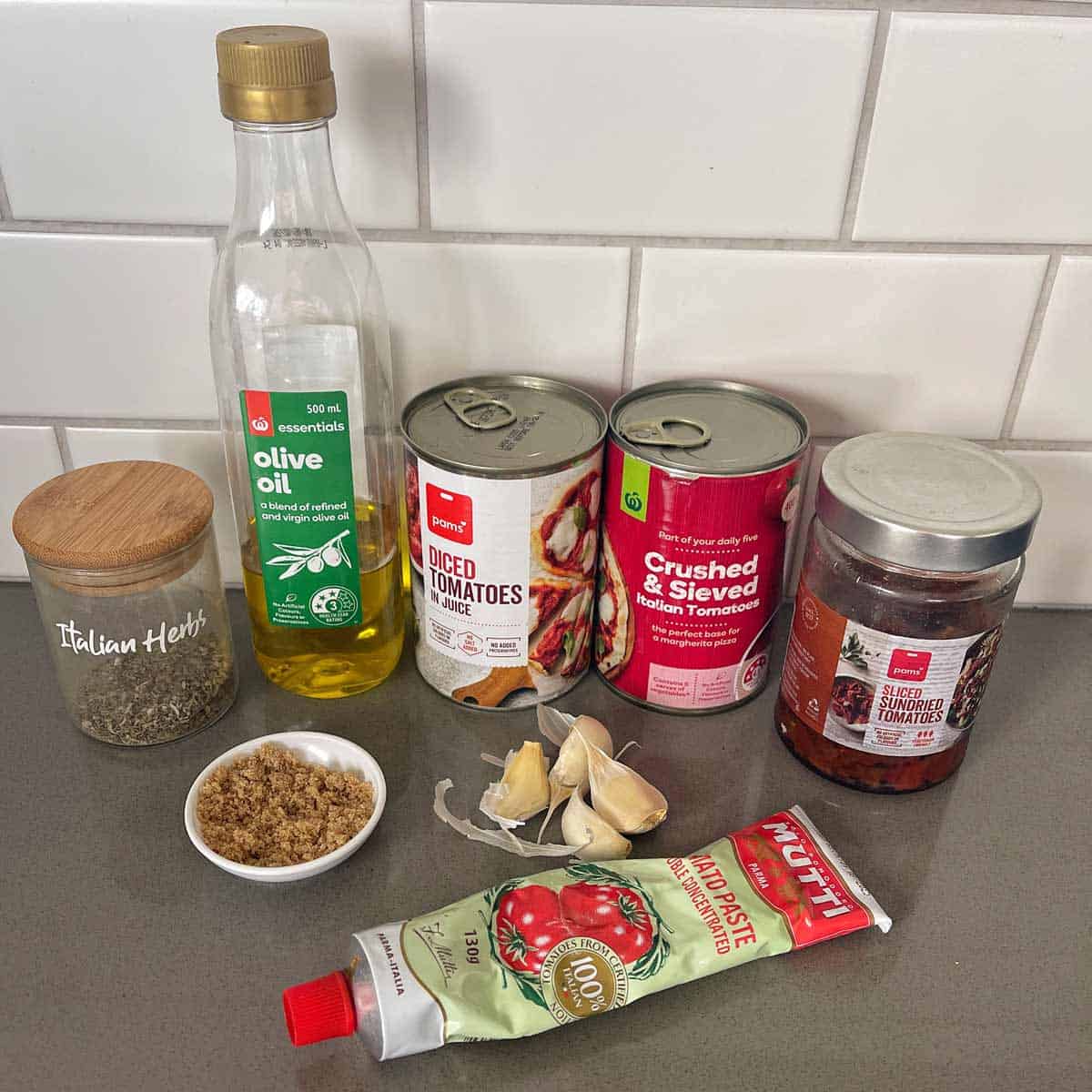 A selection of pasta sauce ingredients on a counter: olive oil, Italian herbs, brown sugar, garlic, diced tomatoes, crushed tomatoes, sun-dried tomatoes, and tomato paste.
